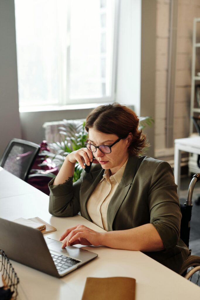 A person using a wheelchair at a desk working on a laptop and taking a phone call.
