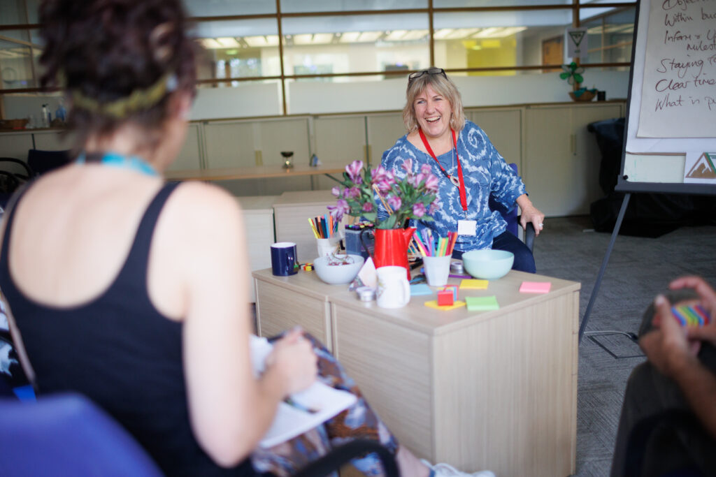 A smiling woman sits at a table, engaging in positive communication with a group during a discussion in an open plan office.