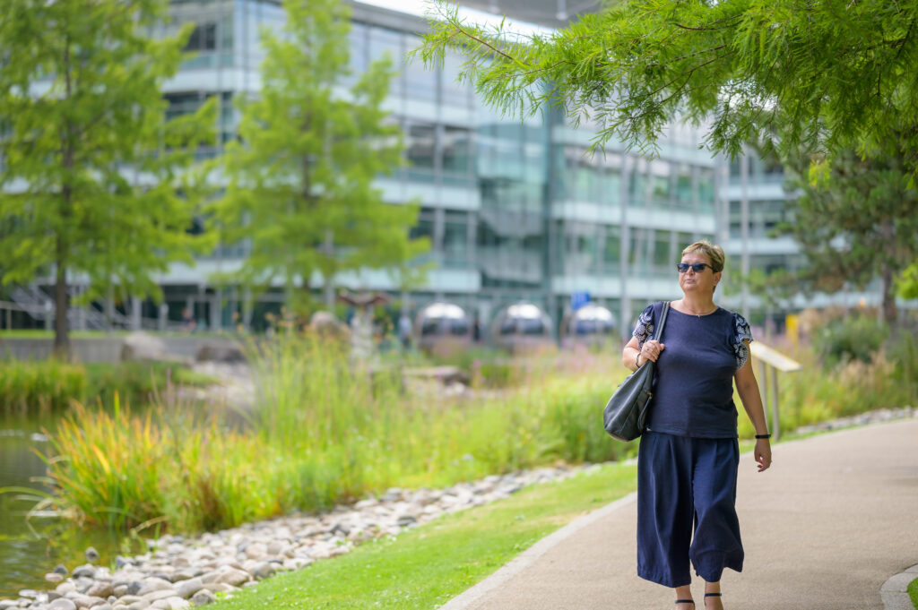 A woman wearing sunglasses walks along a scenic path near a modern office building, carrying a bag and heading to work.