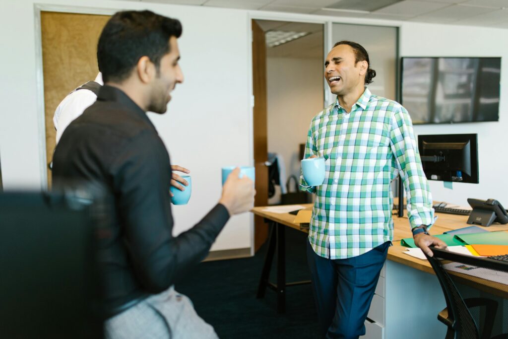 A group of male colleagues standing in an office holding mugs while they talk to each other and laugh.